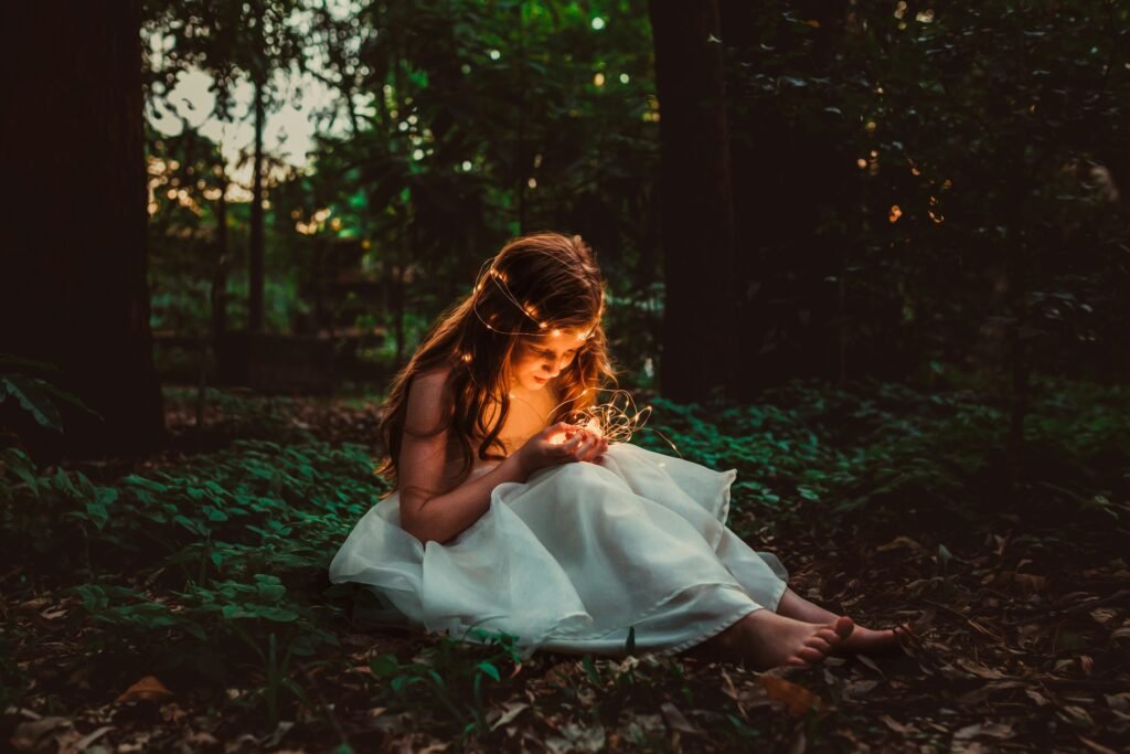 A young girl in a white dress sits barefoot in a forest, illuminated by string lights, creating a magical atmosphere.